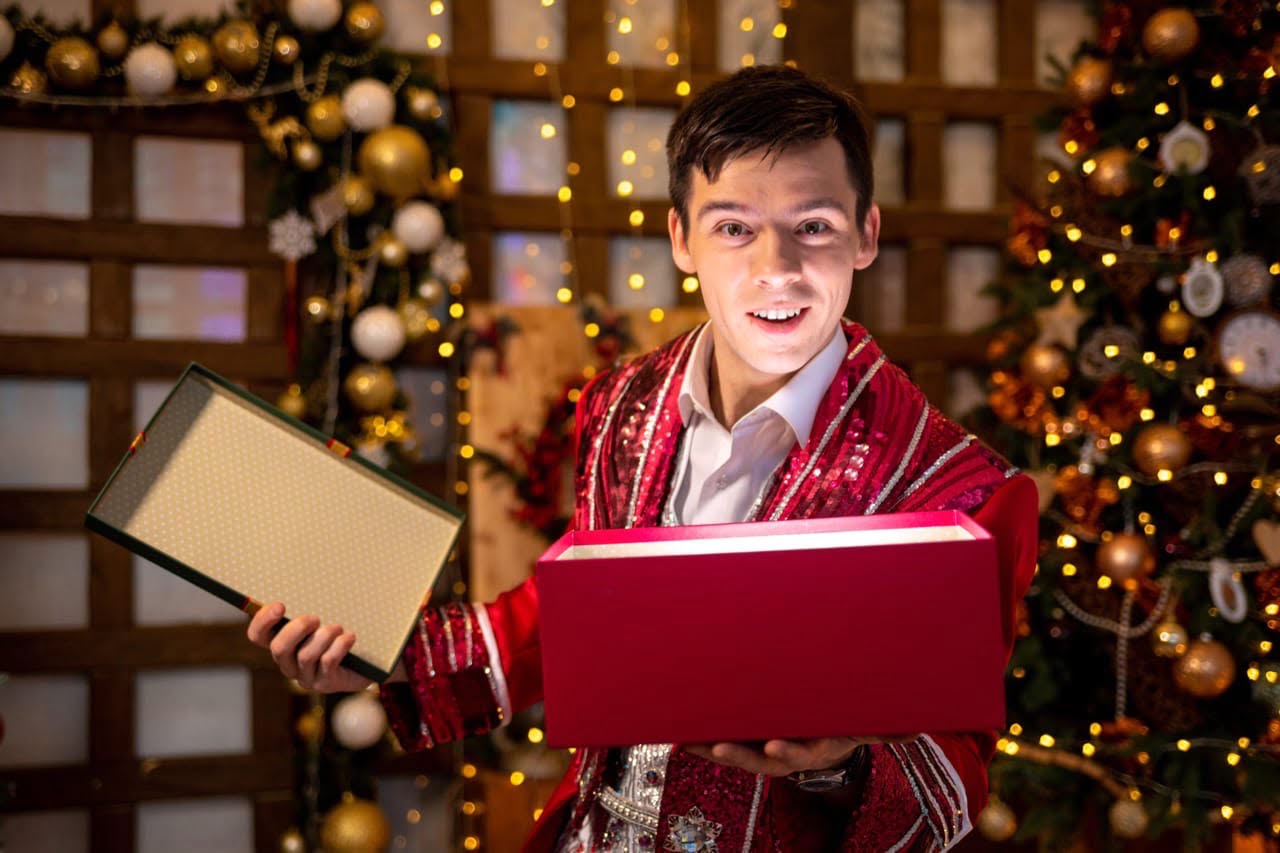 Фокусник в красном фраке с волшебной коробкой на фоне новогодней елки